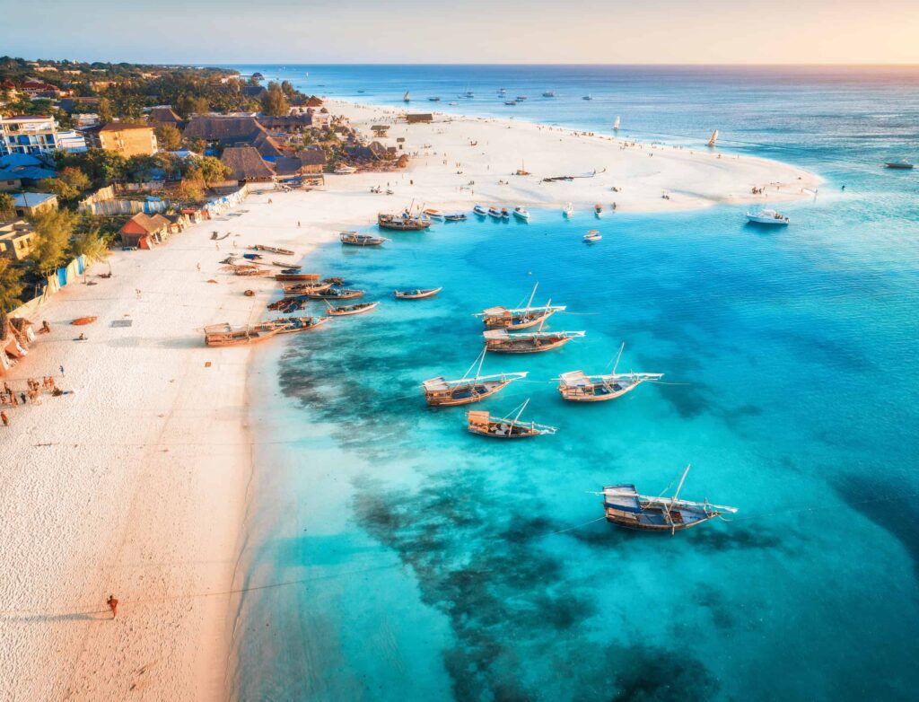 Aerial view of the fishing boats on tropical sea coast with sandy beach at sunset. Summer holiday on Indian Ocean, Zanzibar, Africa. Landscape with boat, buildings, transparent blue water. Top view